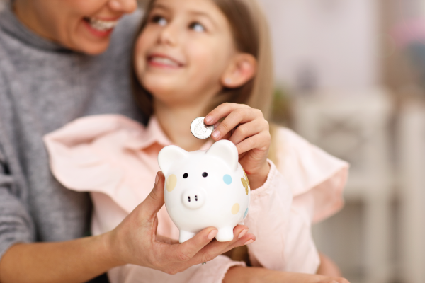 Young girl and her mother with a piggybank sitting on a table and inserting coins into the piggybank